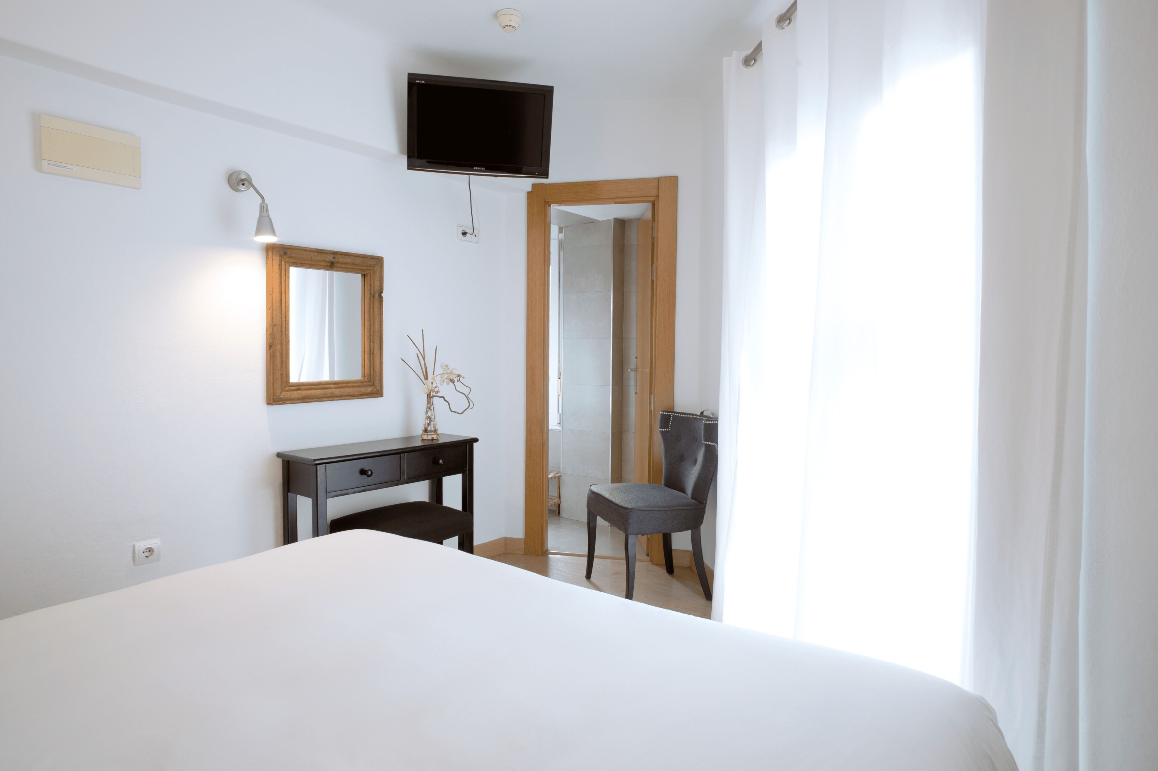 A bedroom with a bed, a desk, a chair, a TV mounted on the wall, and a window with curtains.