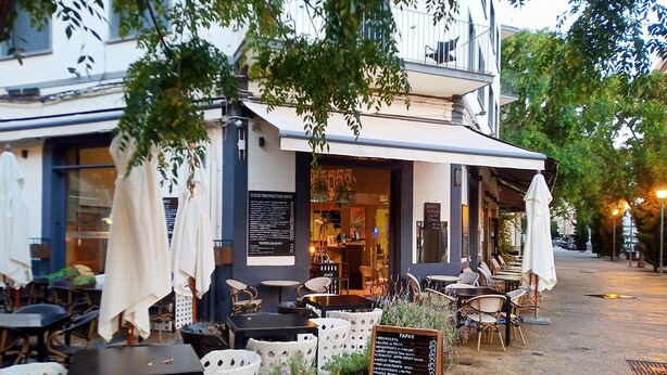 An outdoor cafe with tables and chairs, a menu board, and a building with a balcony.