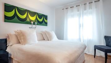 A neatly made bed with white linens, a modern abstract painting on the wall, and a chair by the window.