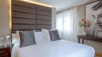 A modern hotel room with a large bed, a bedside table with a lamp, a decorative vase with flowers, and a framed portrait on the wall.
