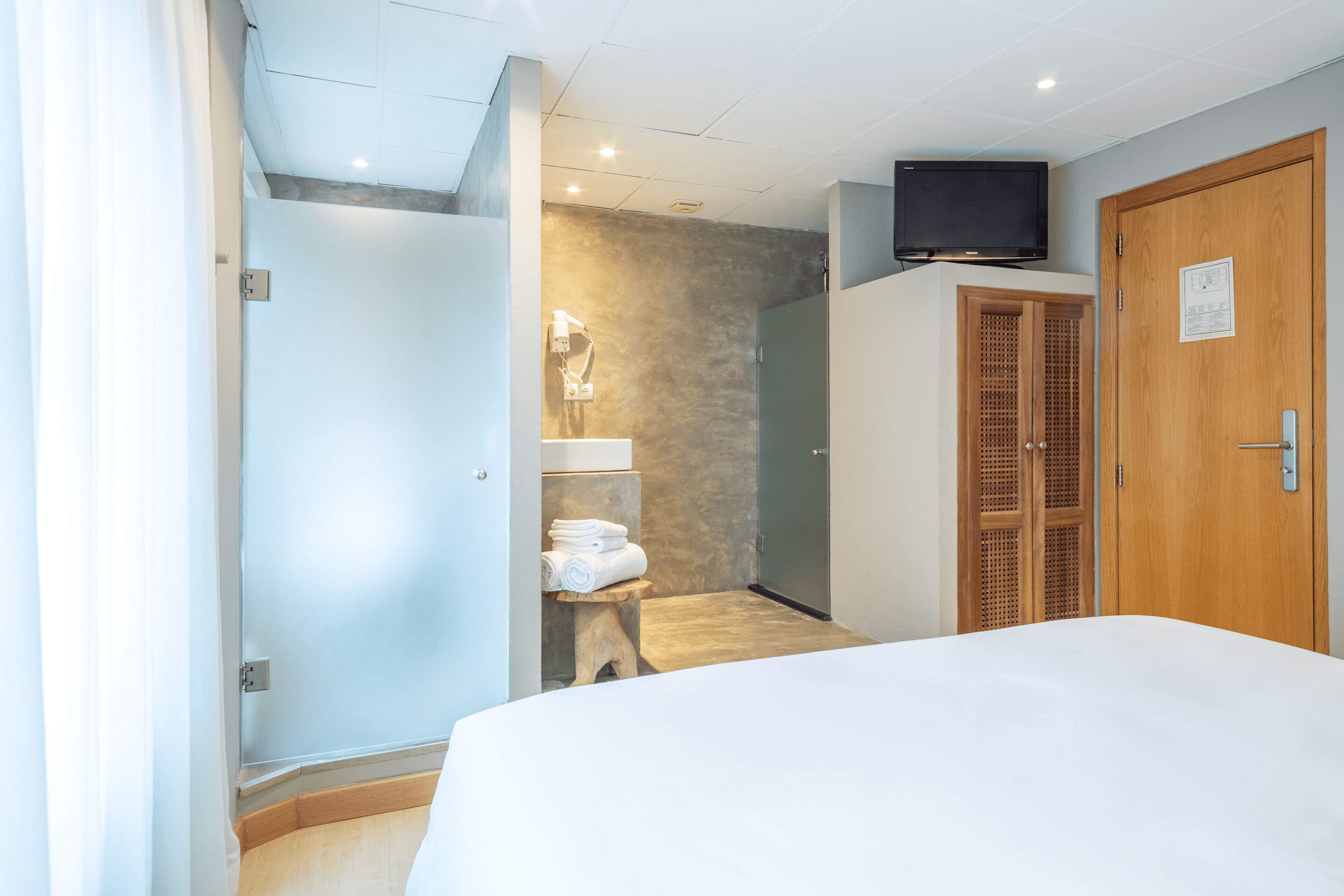 Ett hotellrum med en säng, en garderob i trä, en tv och ett badrum med en handduksställning.