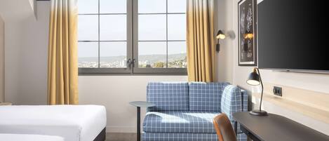 A hotel room with two beds, a sofa, a desk, and a view of the city.