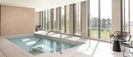 A modern indoor swimming pool with clear glass walls, a sunlit area with a table and chairs, and a view of greenery outside.