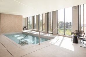 A modern indoor swimming pool with clear glass walls, a sunlit area with a table and chairs, and a view of greenery outside.