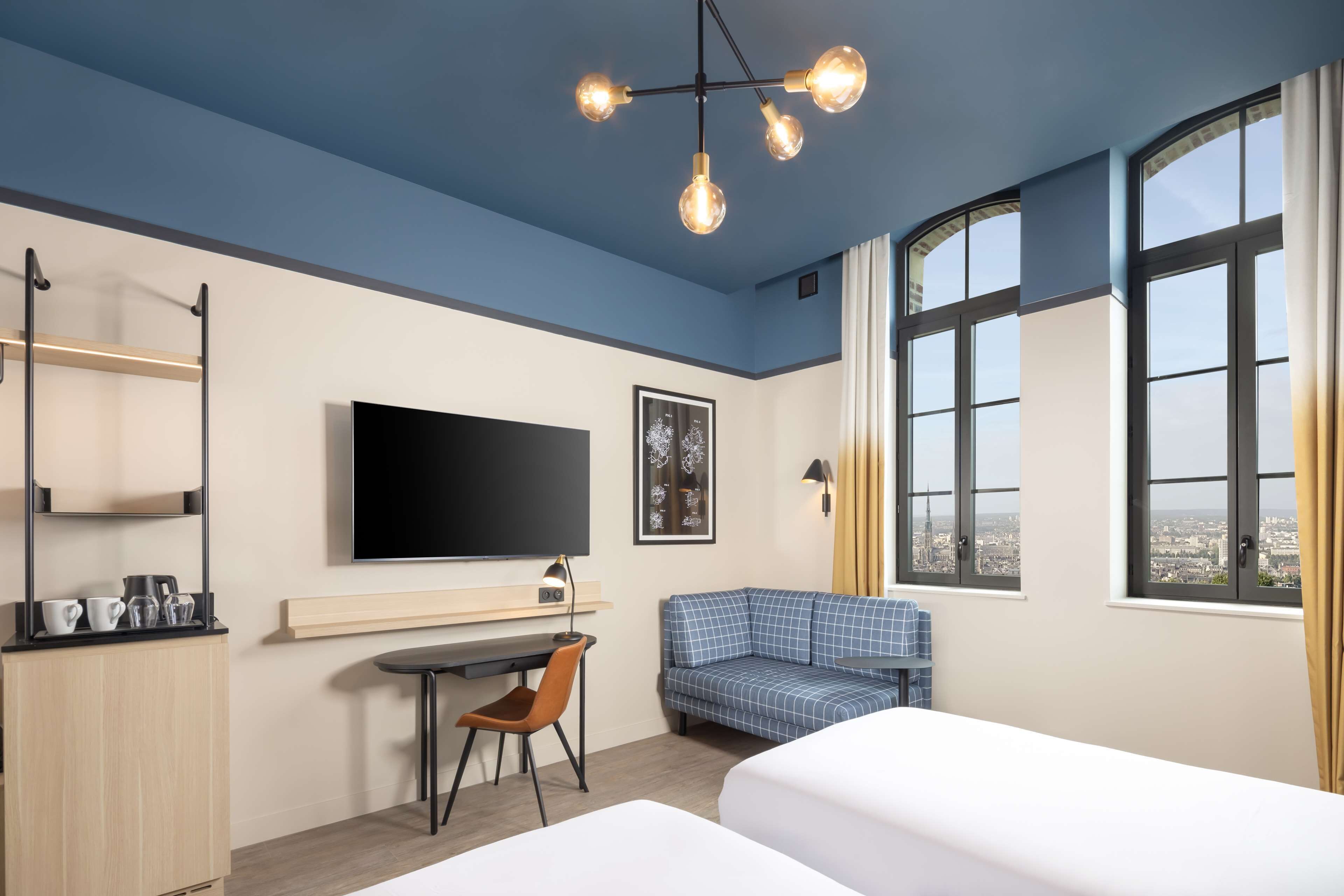 Moderni hotellihuone, jossa on suuri sänky, työpöytä tuolilla, televisio ja suuret ikkunat, joista avautuu näkymä kaupunkiin.