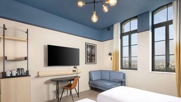 Moderni hotellihuone, jossa on suuri sänky, työpöytä tuolilla, televisio ja suuret ikkunat, joista avautuu näkymä kaupunkiin.