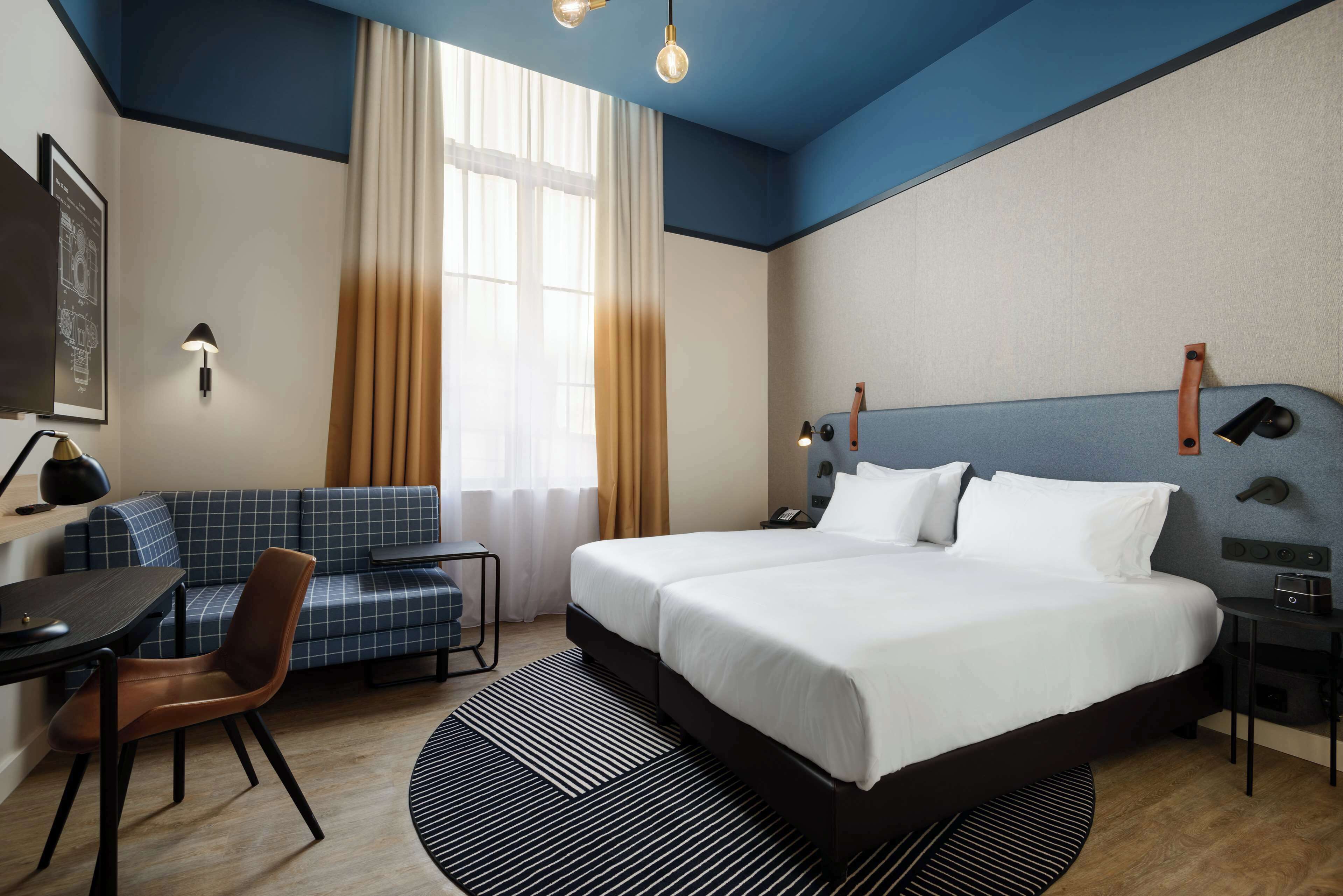 A hotel room with a large bed, a desk with a chair, a sofa, and a window with curtains.