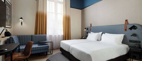 A hotel room with a large bed, a desk with a chair, a sofa, and a window with curtains.