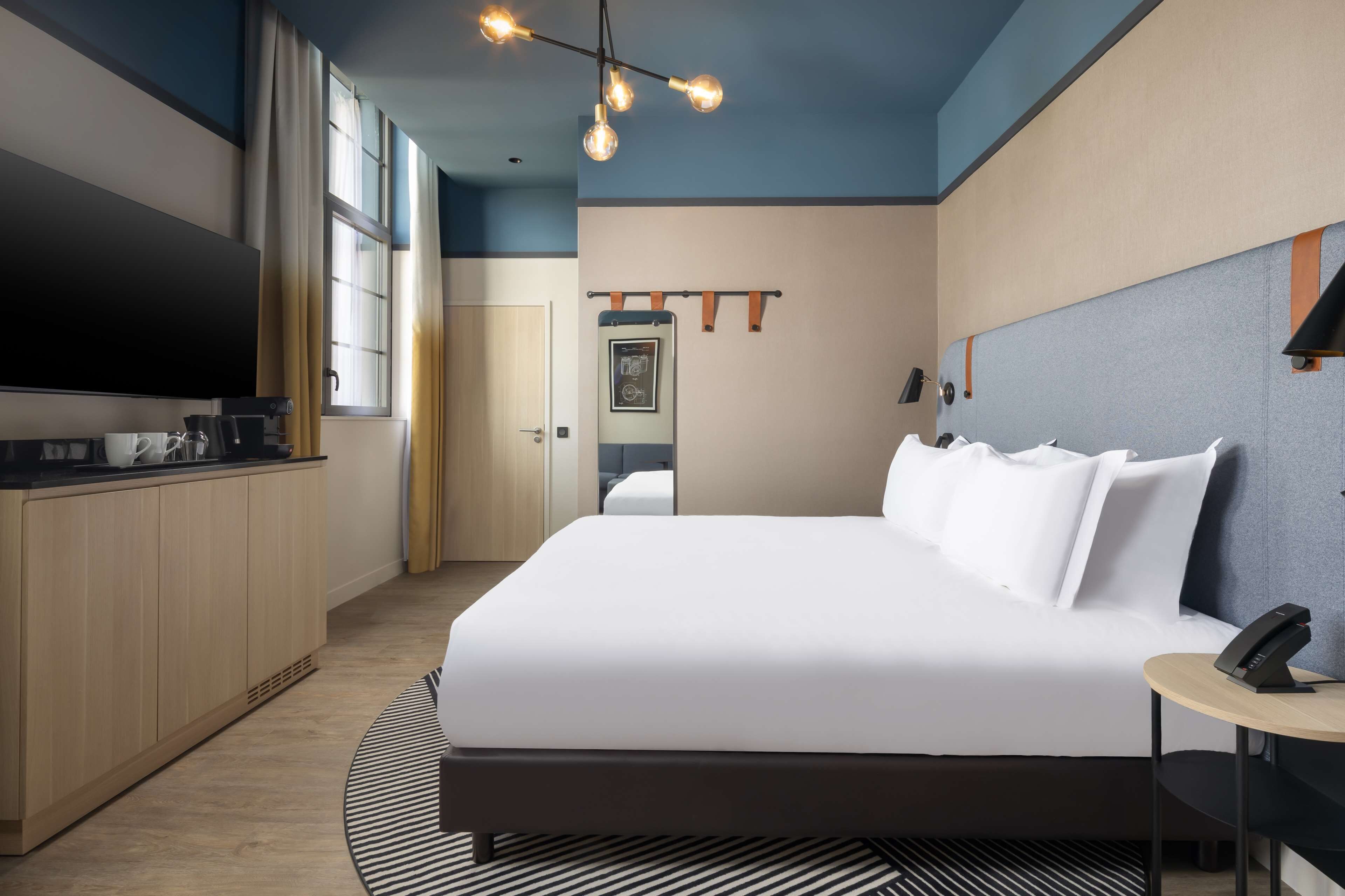 Una habitación de hotel moderna con una cama grande, un televisor de pantalla plana, un escritorio con teléfono y una lámpara de techo original.