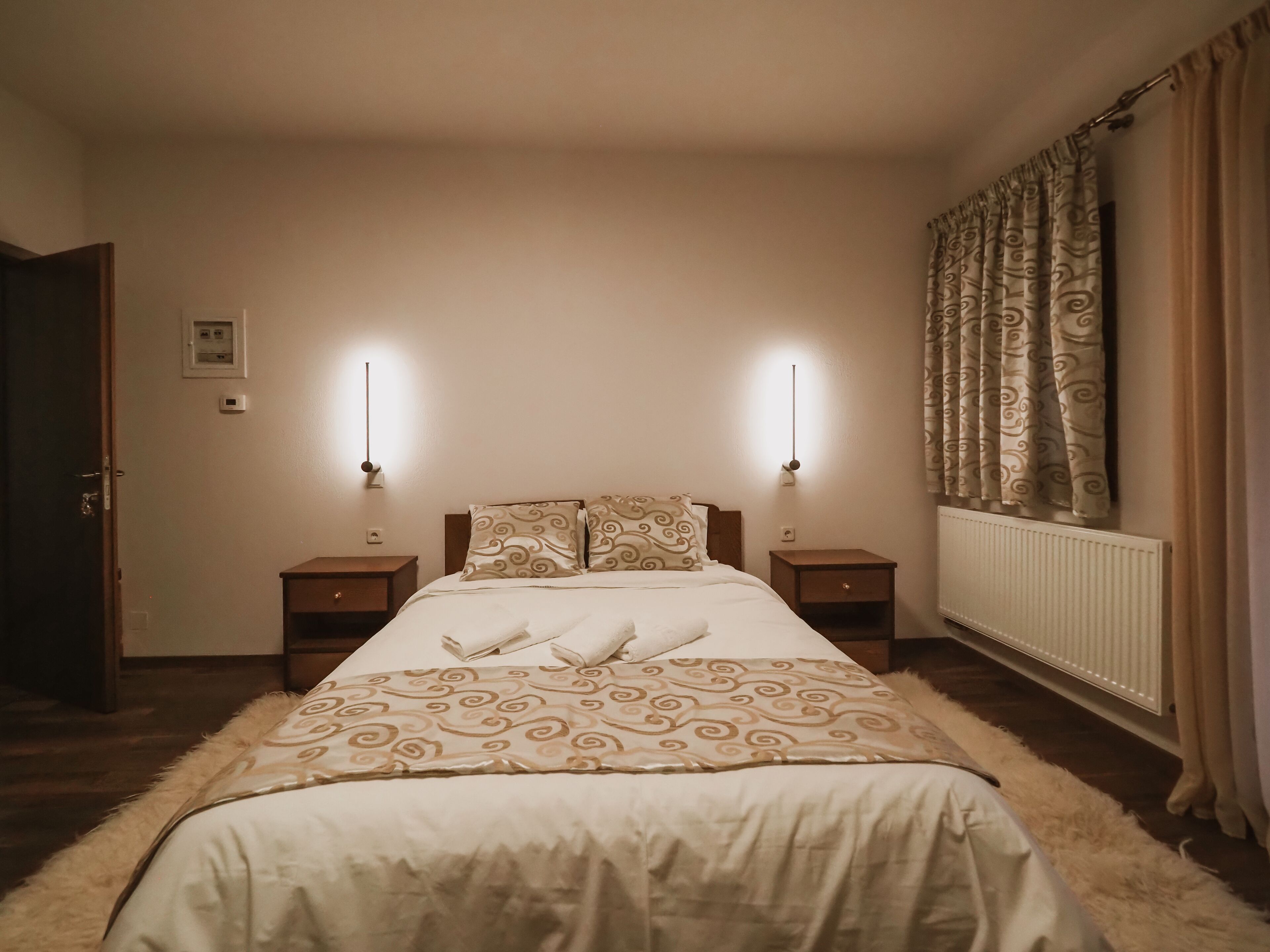 Un dormitorio con cama, dos mesitas de noche, un radiador y una puerta, además de una lámpara de pared.