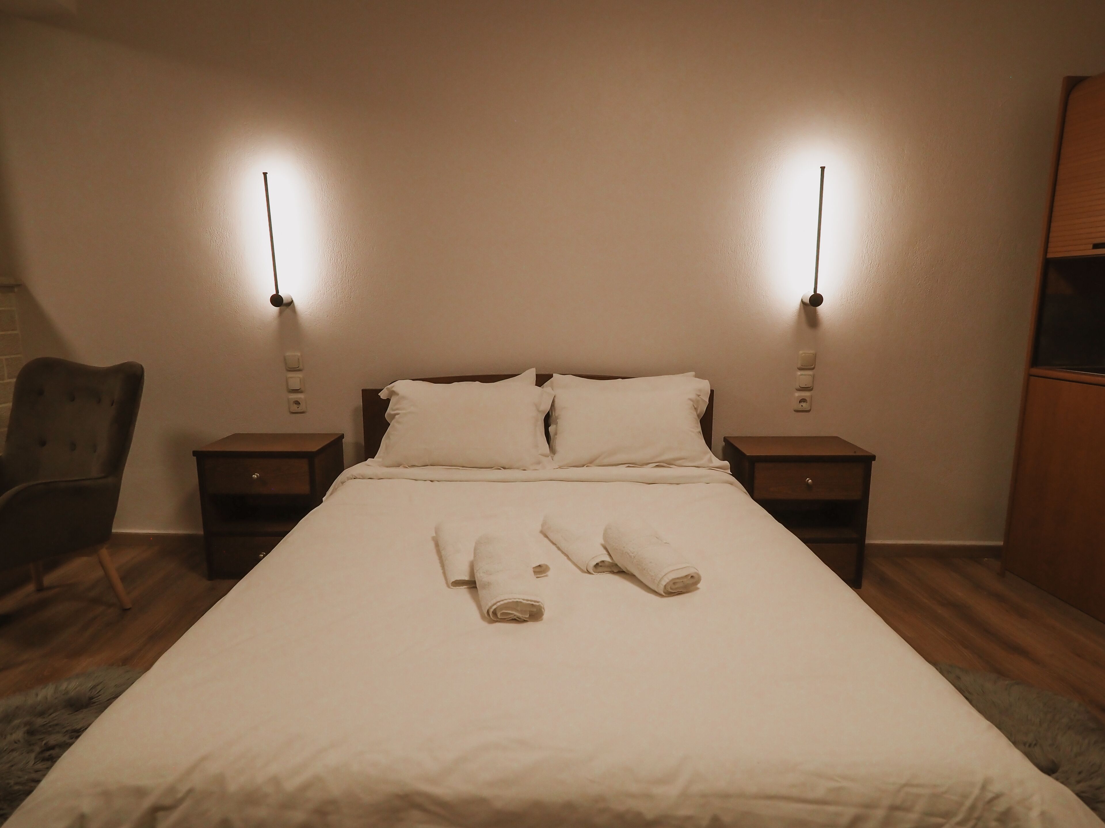 A neatly made bed with white linens and two rolled towels on the bed. There are two bedside tables with lamps on either side of the bed.