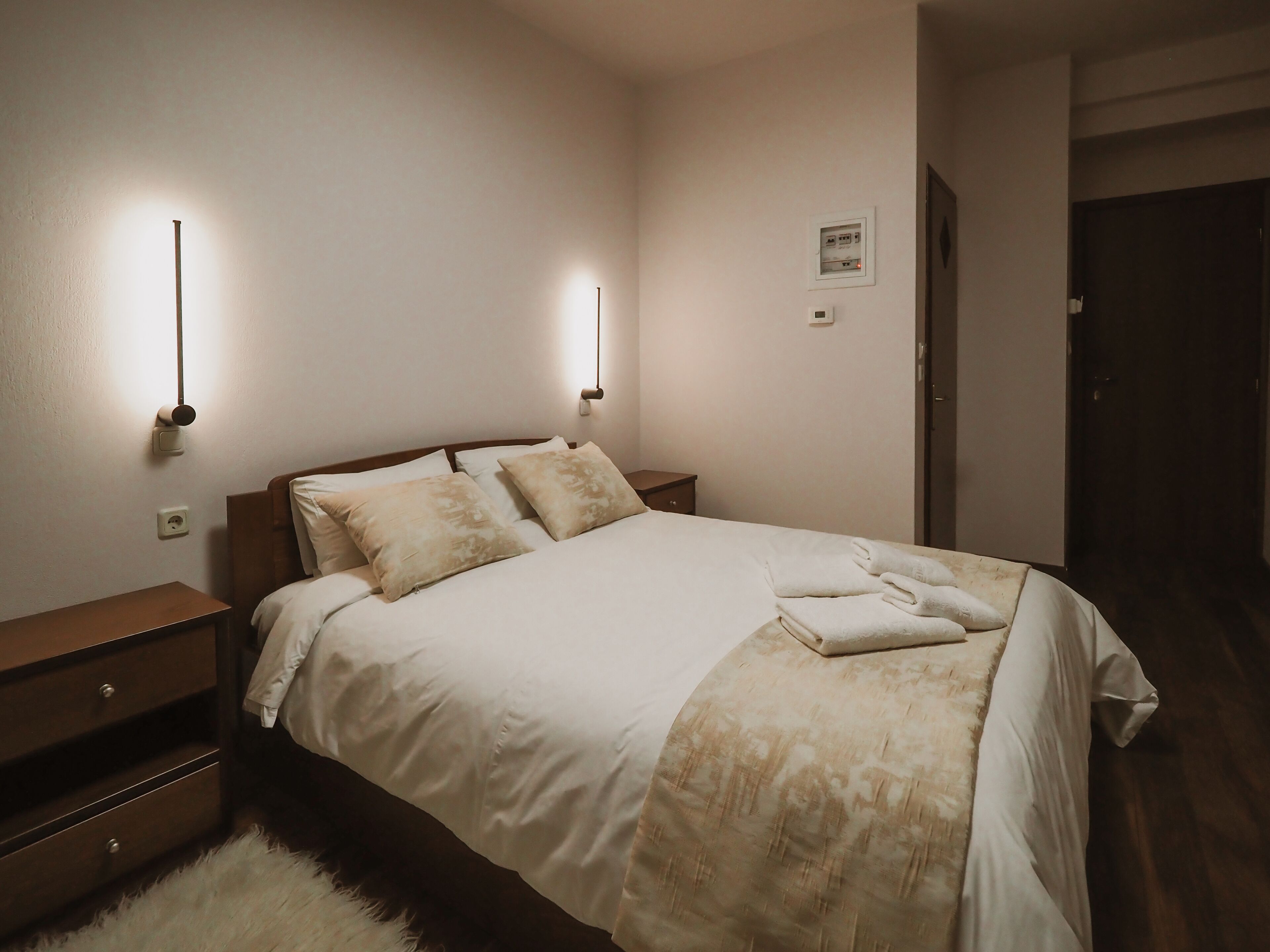 A neatly made bed with a white and beige duvet, a wooden nightstand with a lamp, and a small bedside table.