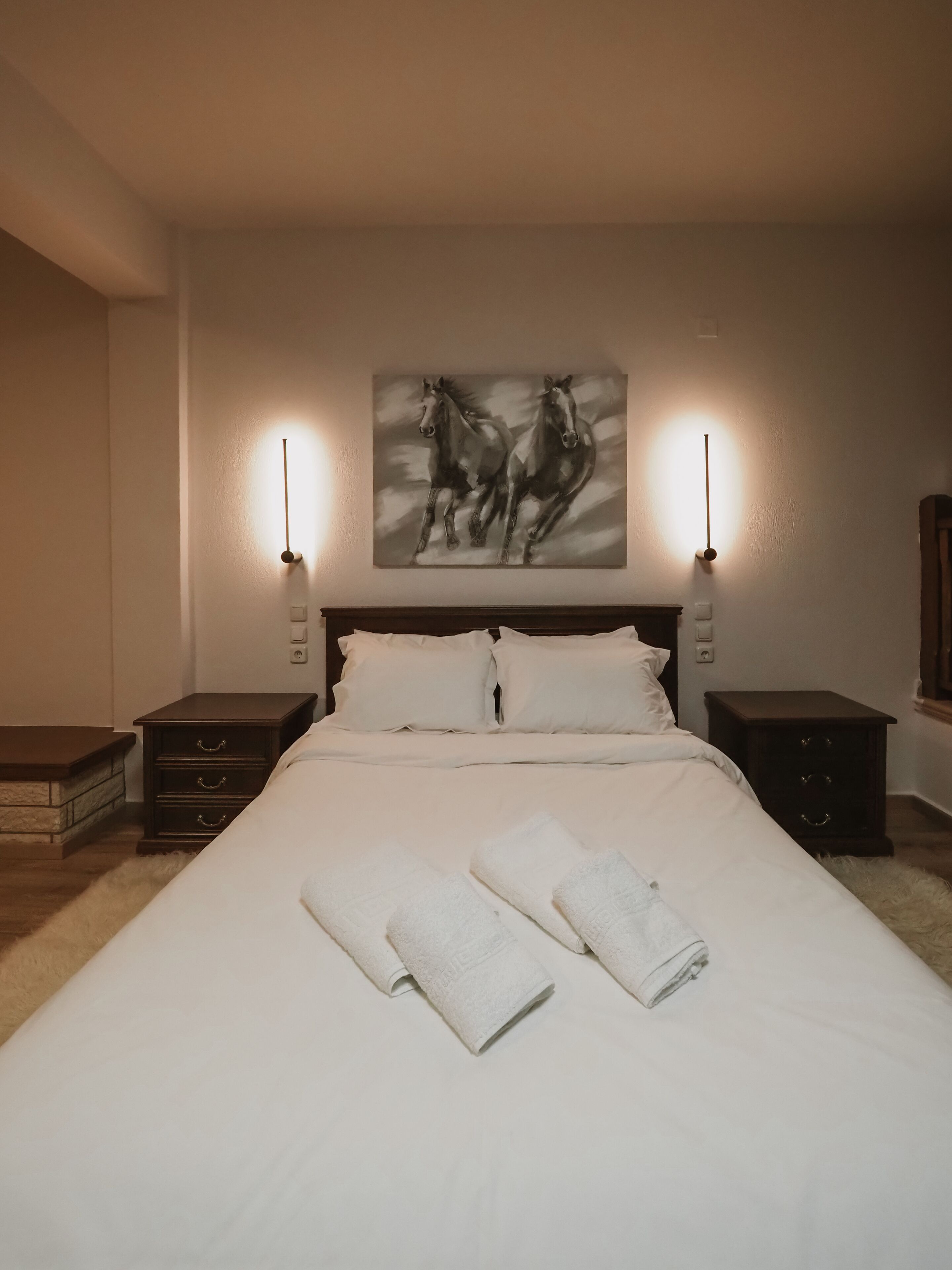 Una cama bien hecha con sábanas blancas y dos toallas, flanqueada por dos mesitas de noche con lámparas, en una habitación con un cuadro grande de caballos en la pared.
