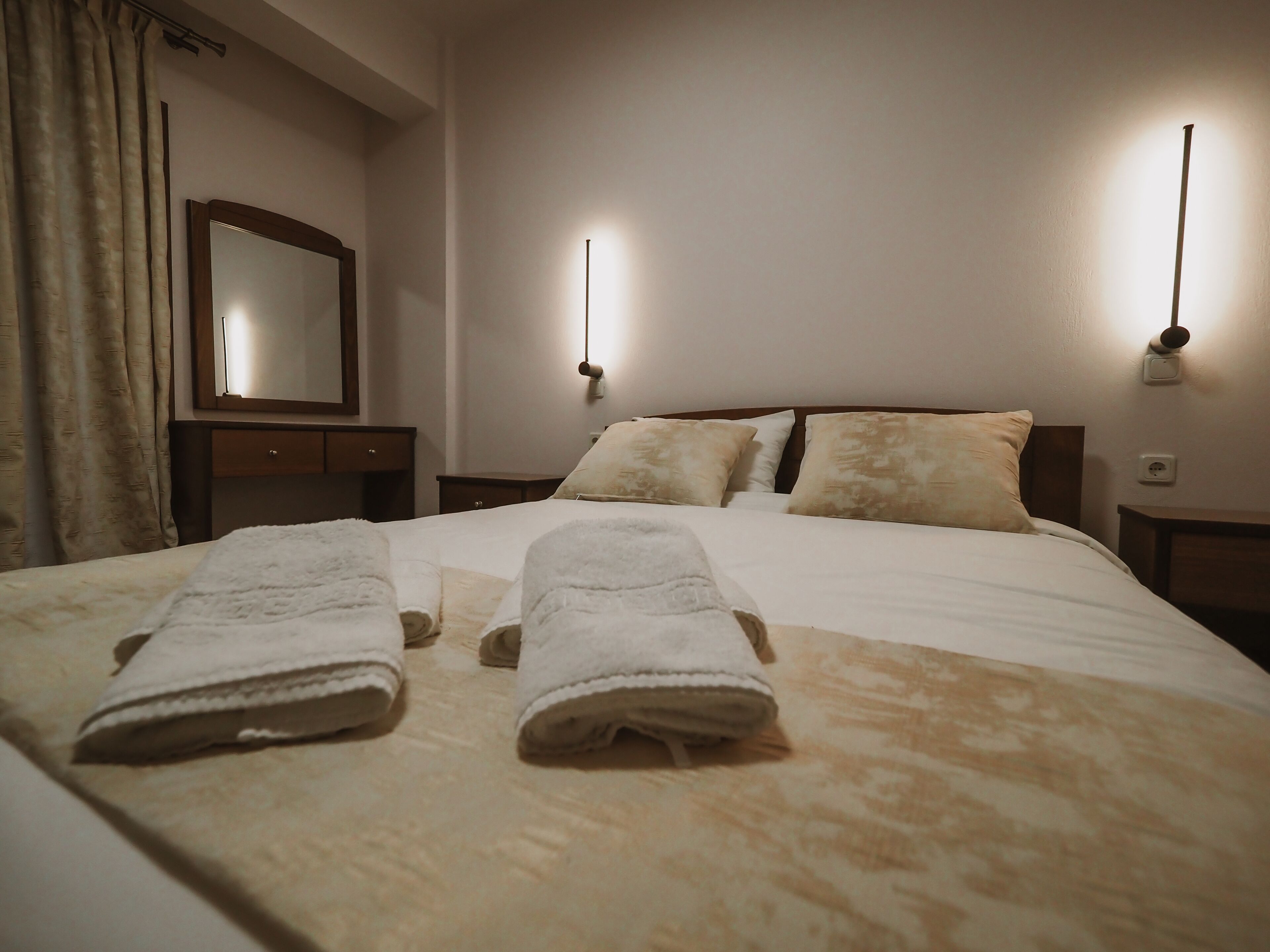 A hotel room with a neatly made bed, two bedside lamps, a mirror, and a wooden nightstand.
