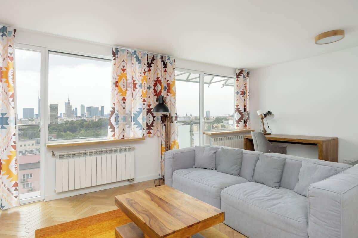 Ein modernes Wohnzimmer mit einem grauen Sofa, einem Holztisch und Blick auf eine Stadtlandschaft durch große Fenster.
