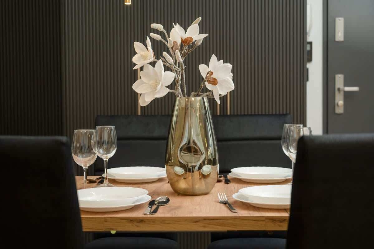 A dining table set with white plates, glasses, and cutlery, centered with a vase of white flowers.