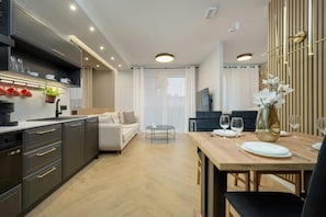 A modern kitchen with dark cabinetry, a dining area with a wooden table, and a living space with a sofa and a TV.