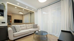 Un salon moderne avec un canapé, une table basse en verre, un téléviseur et une grande fenêtre ornée de rideaux légers.
