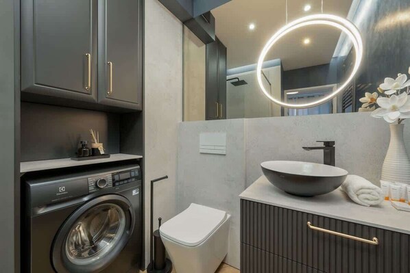 A modern bathroom with a washing machine, toilet, vanity with a sink, and a mirror with a light.