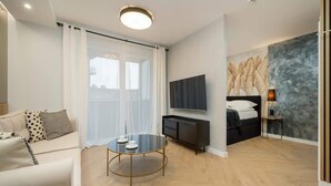 Un salon moderne avec un canapé, une table basse en verre, un téléviseur, un lit et une grande fenêtre avec des rideaux.