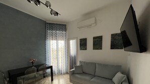 Ein Wohnzimmer mit grauem Sofa, einem Esstisch mit Stühlen, einem an der Wand befestigten Fernseher und einer Klimaanlage.