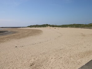 Une vaste plage de sable aux courbes douces, un ciel dégagé et des dunes qui se dessinent à l’horizon.