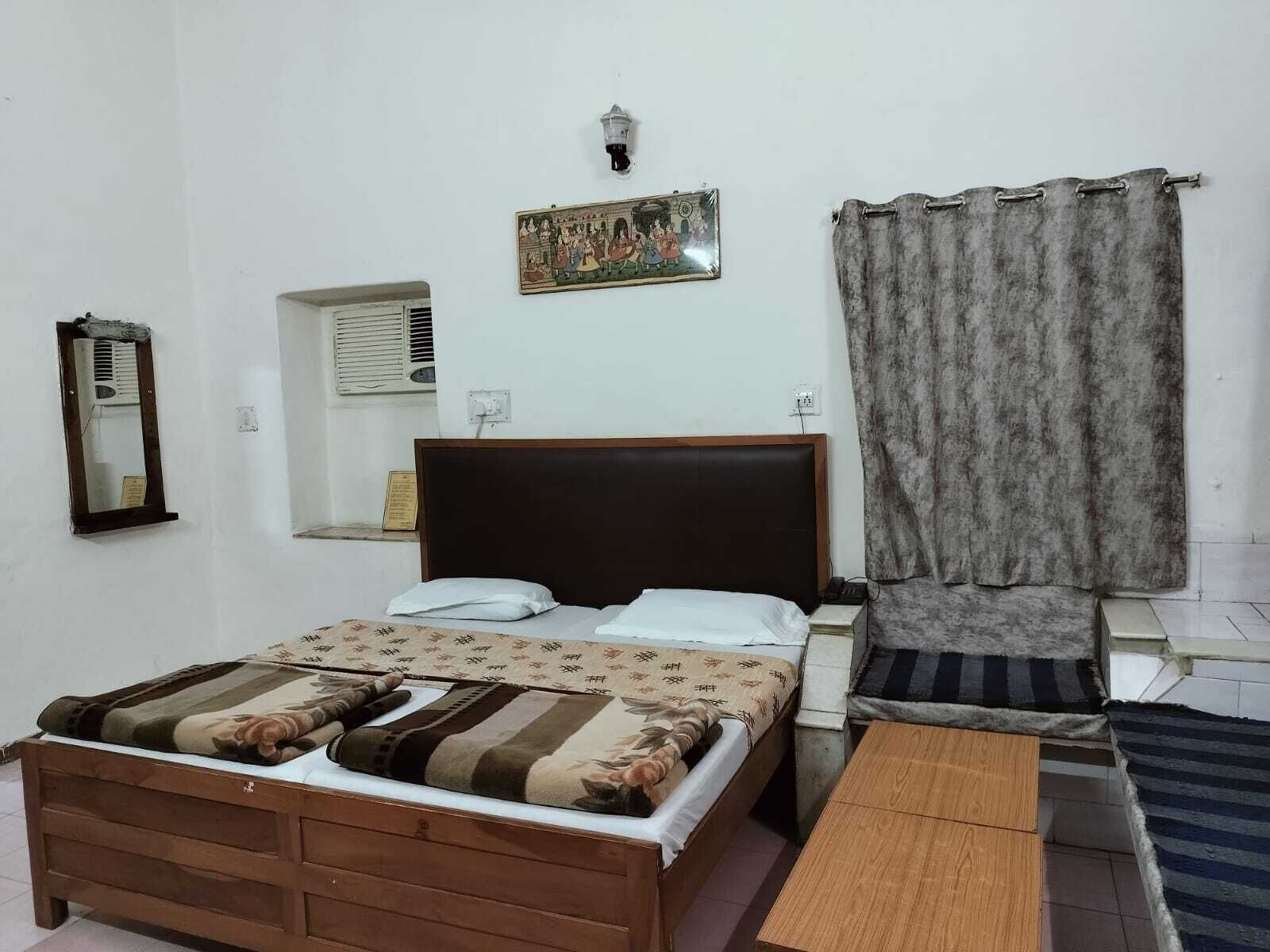 A bedroom with a wooden bed, a nightstand, a small table, a wall-mounted lamp, a window with an air conditioner, and a curtain.