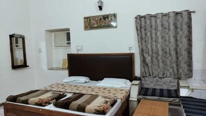 A bedroom with a wooden bed, a nightstand, a small table, a wall-mounted lamp, a window with an air conditioner, and a curtain. - Hotel Rawat (Jodhpur)