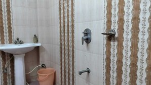 A bathroom with a pedestal sink, a shower with a wall-mounted faucet, and a bucket on the floor. - Hotel Rawat (Jodhpur)
