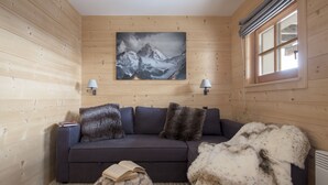Interior de una cabaña de madera con un sofá oscuro, dos lámparas, un cuadro de montañas y una ventana con cortinas.