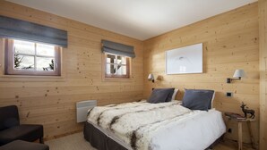 Un dormitorio con paneles de madera, una cama, dos ventanas con persianas, una lámpara de pared y una mesita con una lámpara y un adorno.