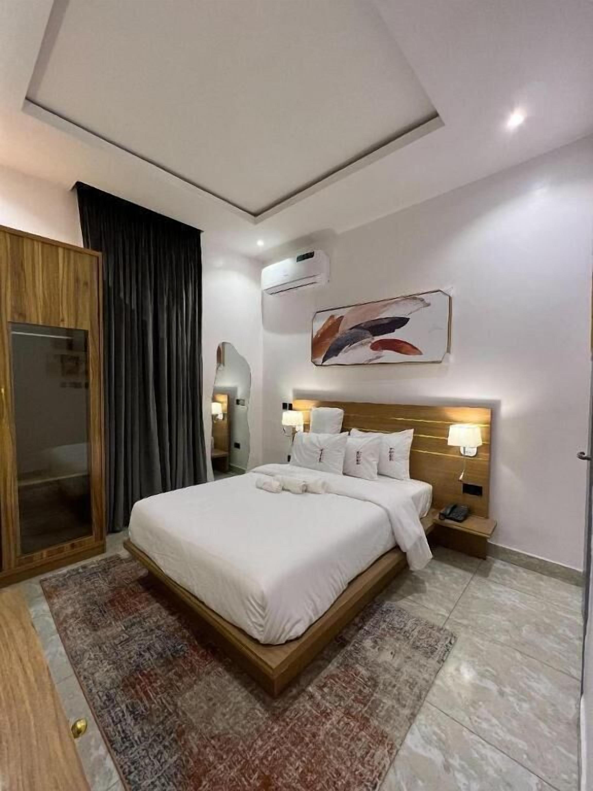 A modern hotel room with a large bed, a wooden wardrobe, a bathroom with a glass door, and a ceiling with recessed lighting.