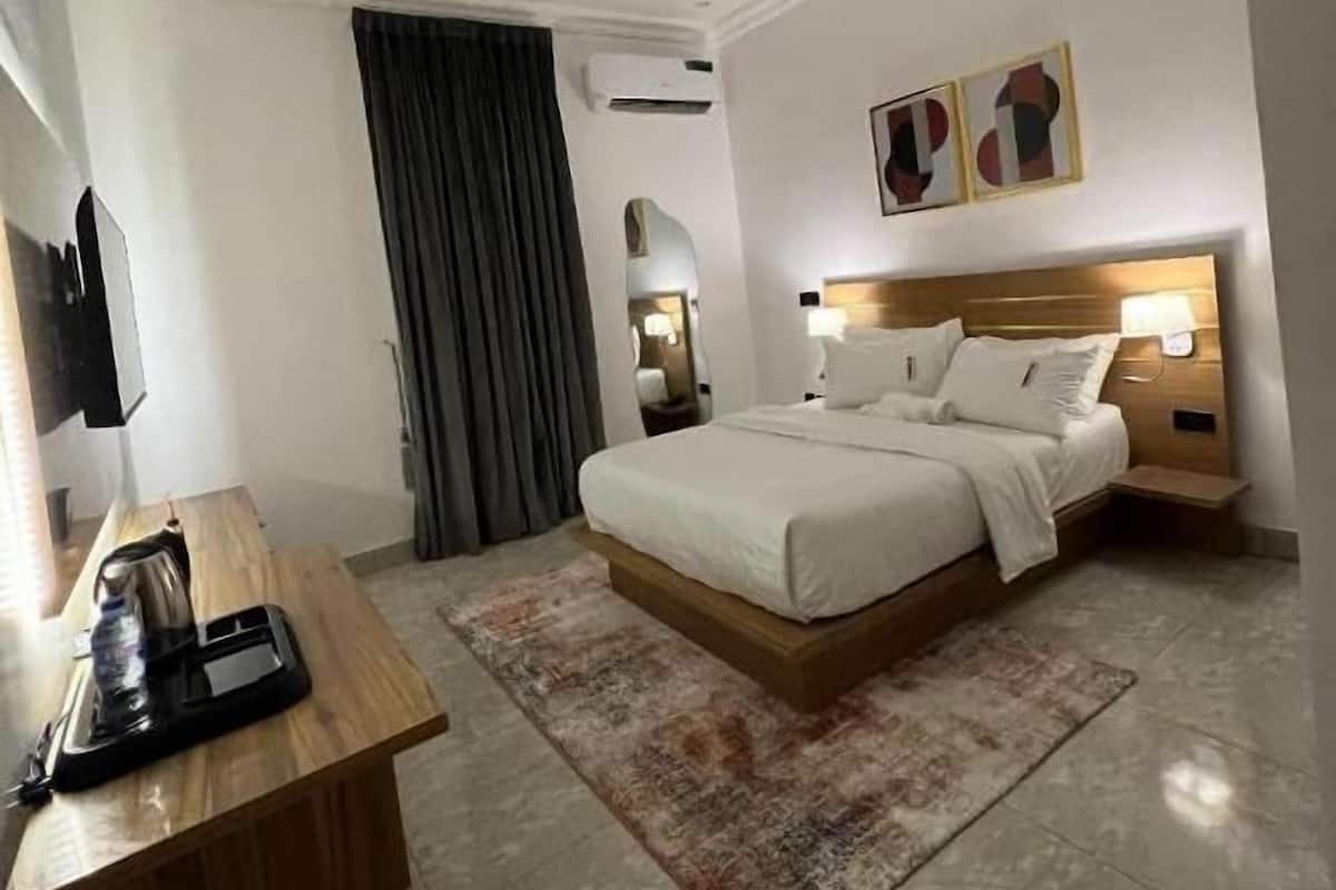 Quarto de hotel com cama, mesas de cabeceira, escrivaninha com chaleira, tapete e unidade de ar condicionado.