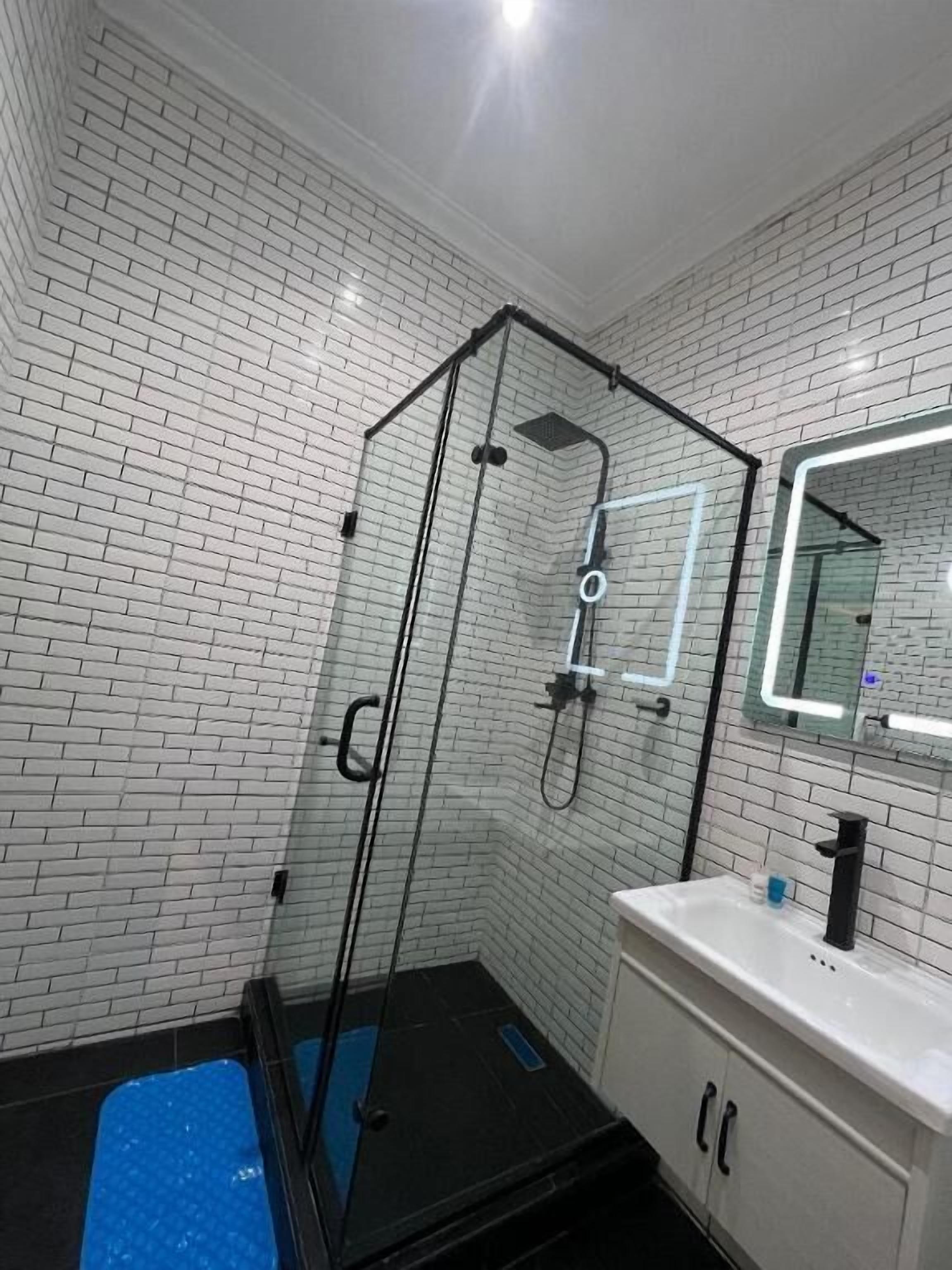 A bathroom with a glass shower enclosure, white tiled walls, a black floor, and a white sink with a mirror above it.