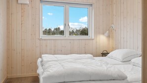Une chambre avec un mur lambrissé en bois, une fenêtre donnant sur des arbres et des immeubles, une lampe de chevet et un lit avec une literie blanche.