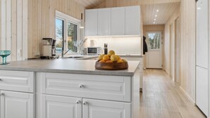 Une cuisine moderne avec des meubles blancs, un grand îlot central agrémenté d’un panier de fruits, et une vue sur l’extérieur grâce aux fenêtres.