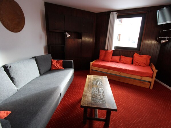 Ein Zimmer mit grauem Sofa, einem hölzernen Bett mit roter Bettwäsche, einem kleinen Holztisch und einem Fenster mit Vorhängen.