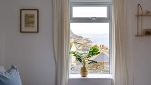 Quarto com uma janela grande, uma cama, uma mesinha de cabeceira, uma planta e um quadro na parede.