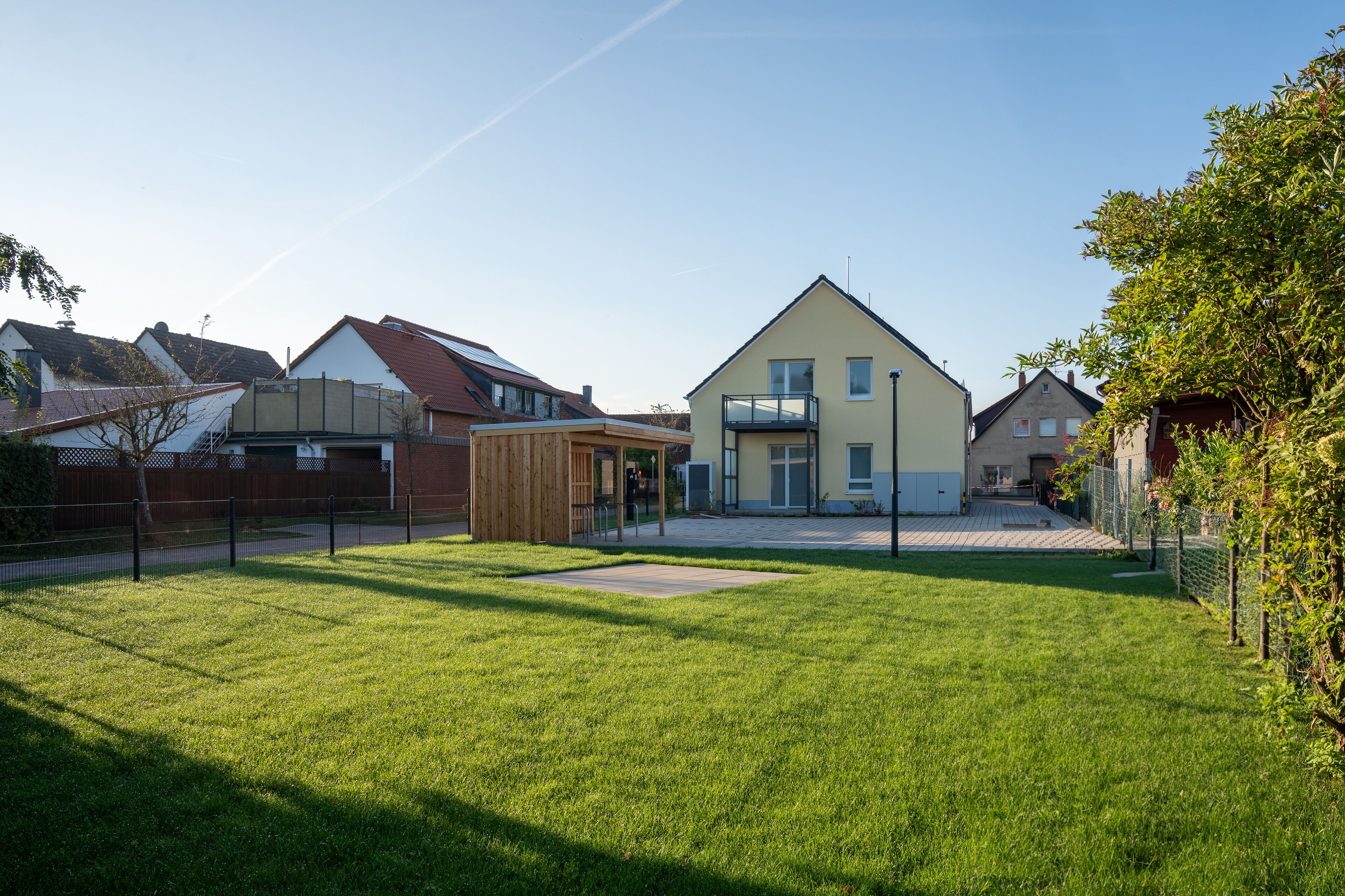 A residential area with a modern house featuring a large front yard, a wooden play structure, and a fenced garden.