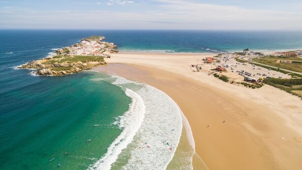 An aerial view of a coastal area with a sandy beach, a rocky outcrop with buildings, and a clear view of the ocean. - Charles Beach House (Ferrel)