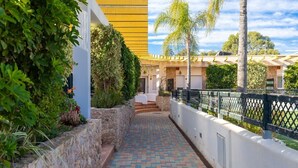 Un chemin menant à un bâtiment à la façade jaune, entouré de verdure et d’un mur de pierre.
