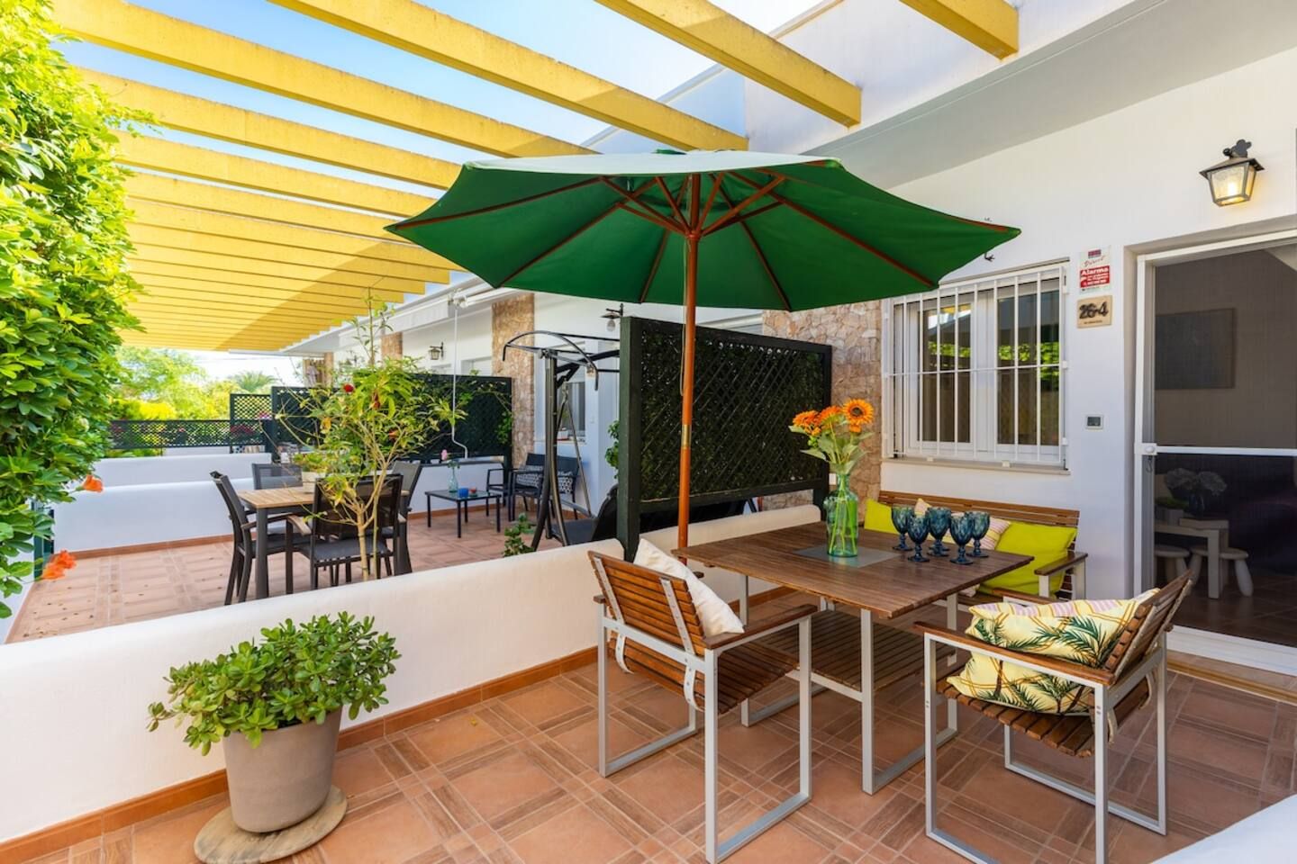 Un patio exterior con una mesa, sillas, un paraguas verde y plantas en macetas.