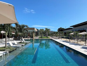 Une piscine entourée de palmiers et de bâtiments modernes, avec des chaises longues et des parasols.