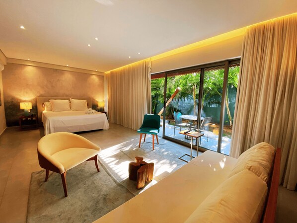 Une chambre d’hôtel moderne dotée d’un grand lit, d’un coin salon avec un fauteuil et une table, et d’un balcon offrant une vue sur la verdure.