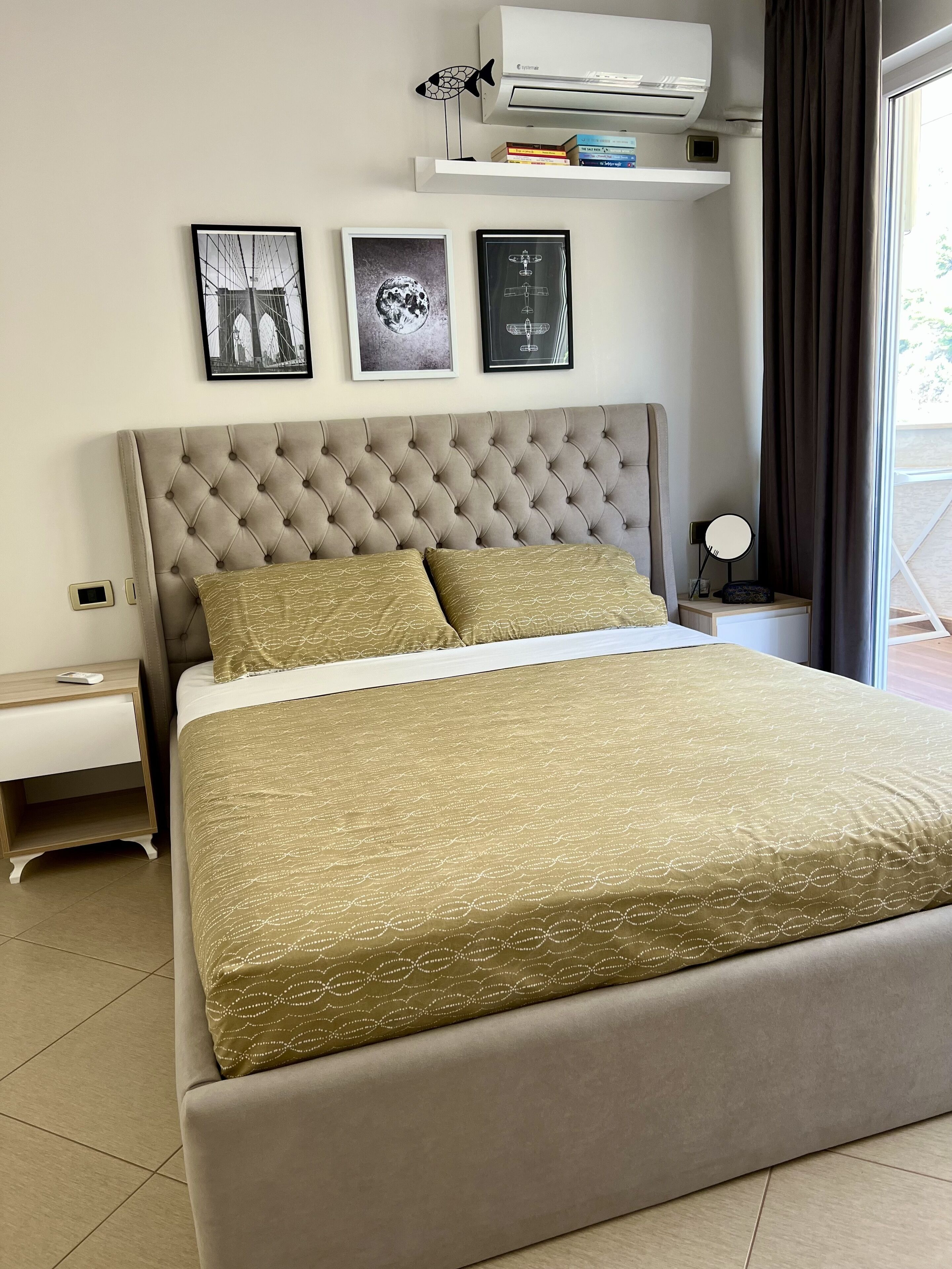 A bedroom with a tufted headboard, a bed with a gold patterned bedspread, a bedside table with a lamp, and a shelf with books and a decorative fish.