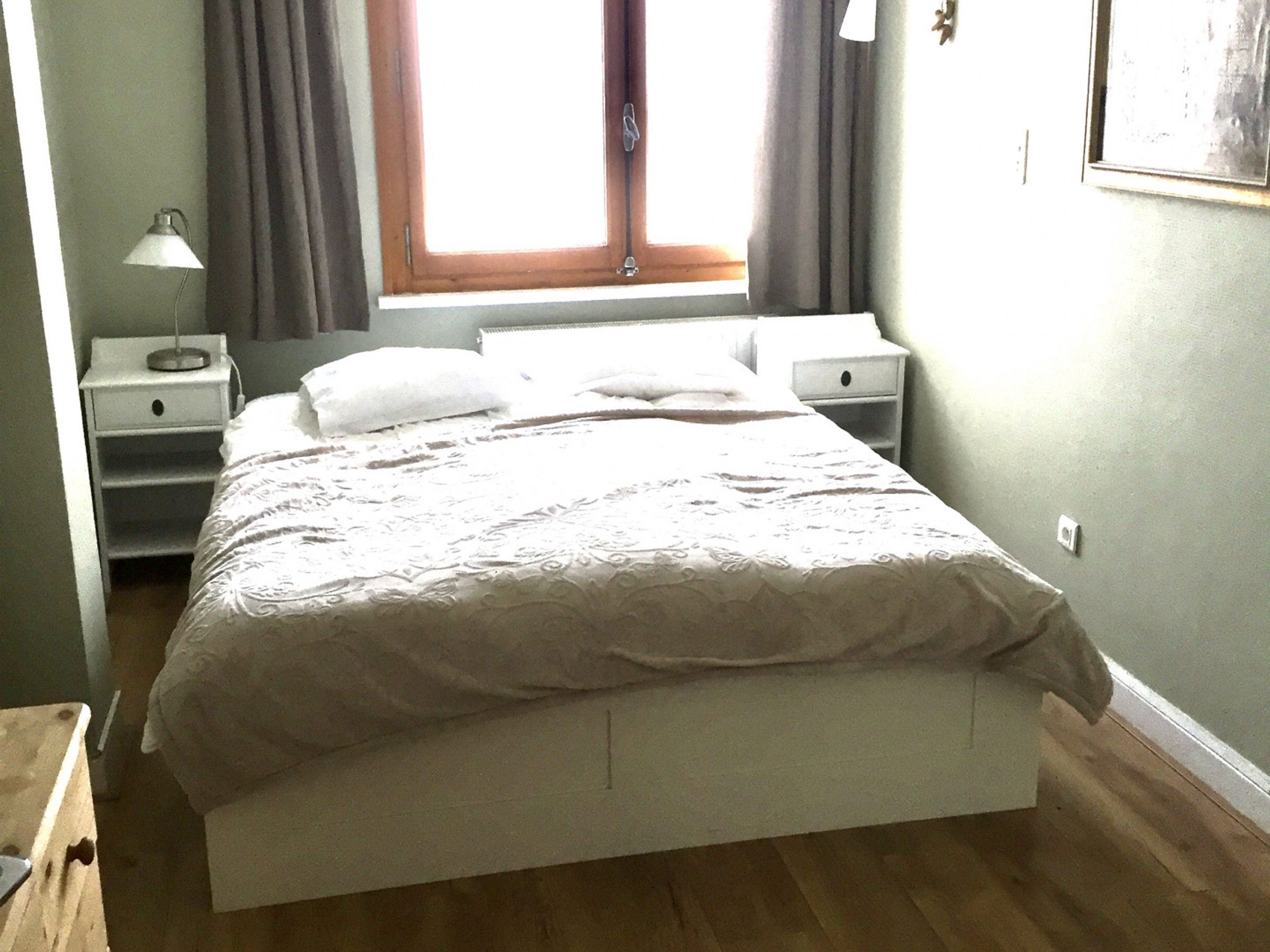 Een bed met een wit hoofdeinde en voeteinde, een nachtkastje en een bijzettafeltje met een lamp.