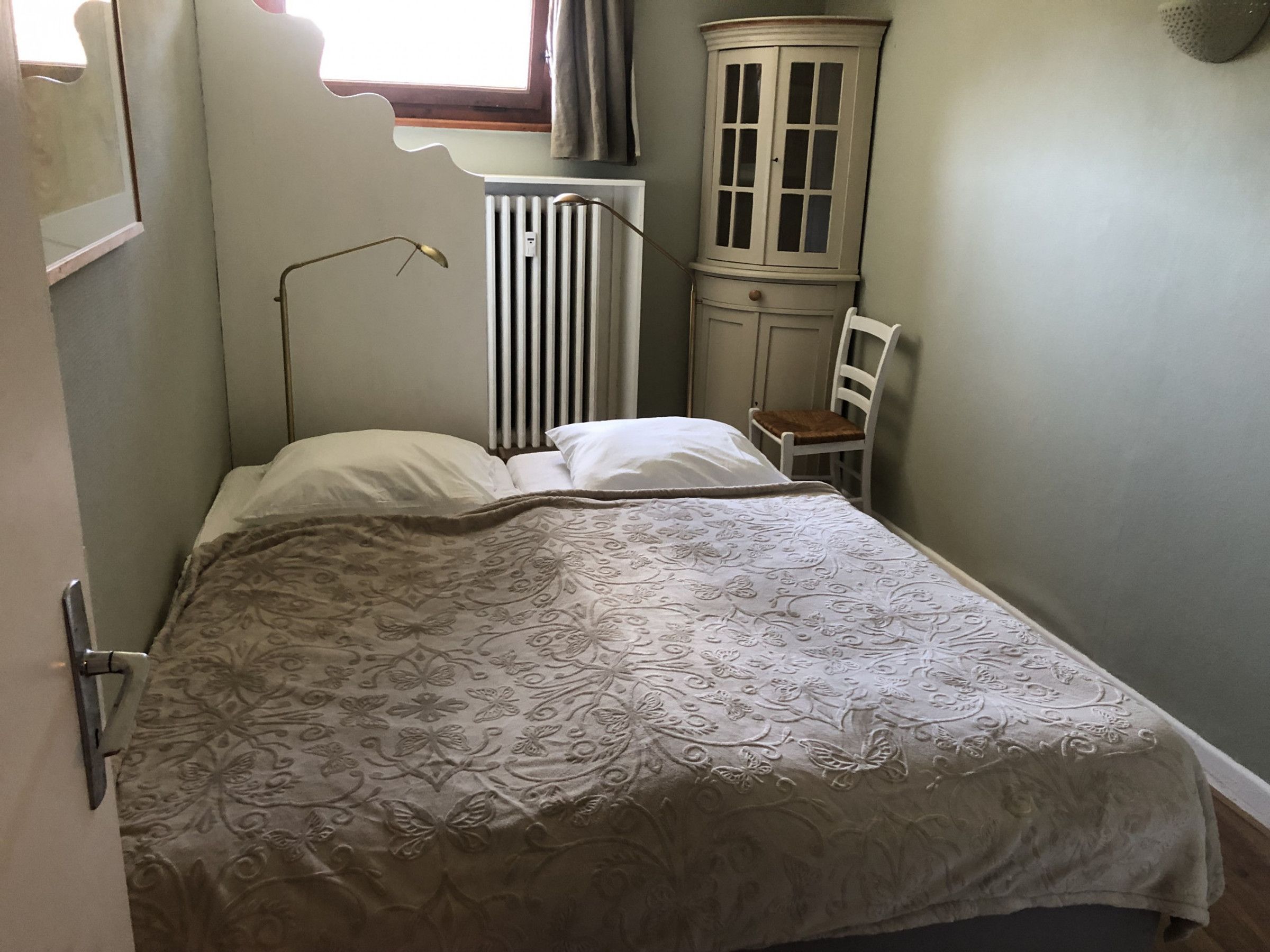 A small, narrow room with a single bed, a radiator, a cabinet, and a chair.