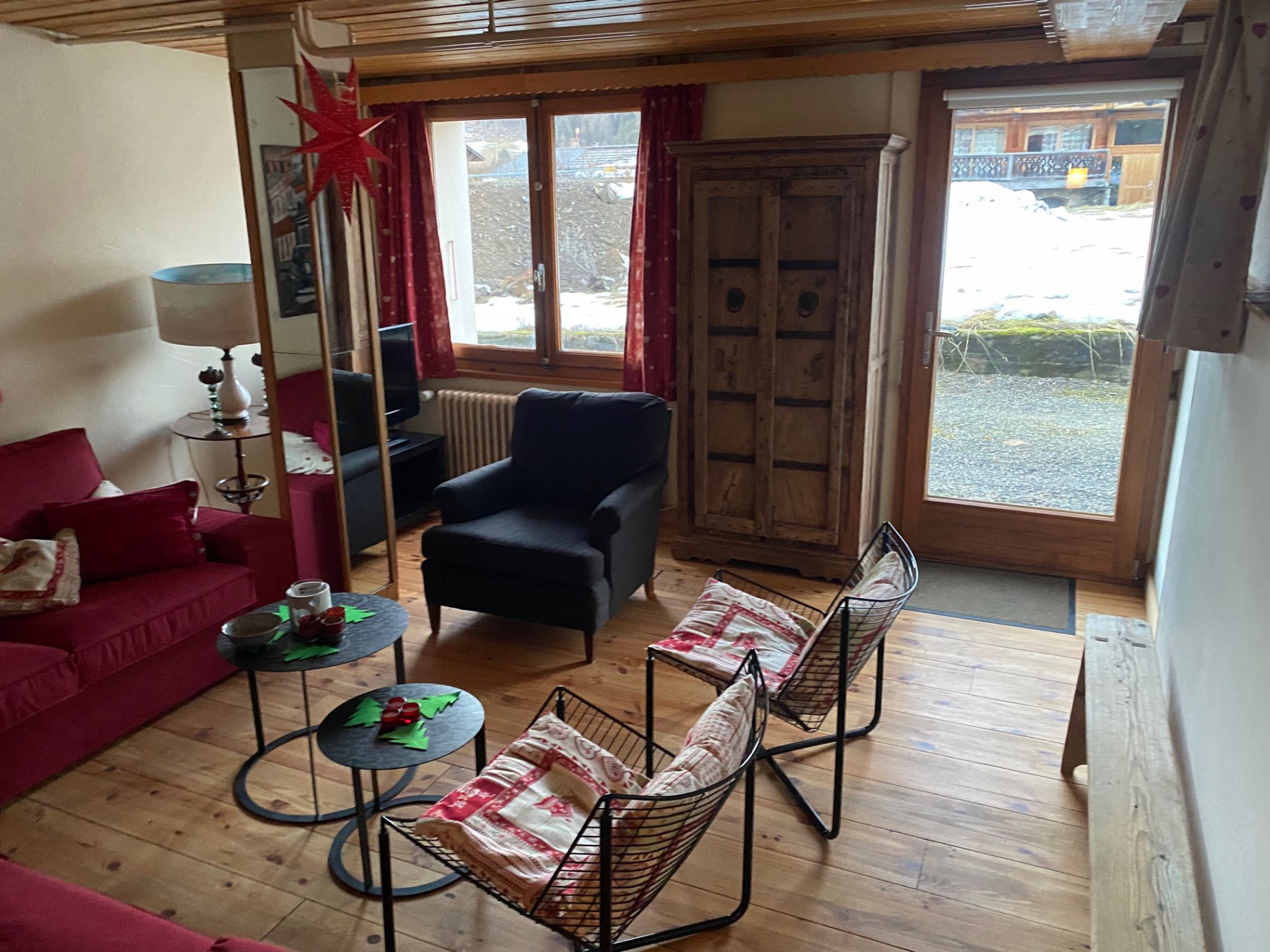 A furnished living room with a red sofa, a black armchair, and two wire-framed chairs. There is a small table with a lamp, a television, and a radiator. A wooden cabinet and a window with curtains are also visible.
