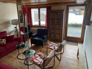 A furnished living room with a red sofa, a black armchair, and two wire-framed chairs. There is a small table with a lamp, a television, and a radiator. A wooden cabinet and a window with curtains are also visible. - Stunning 4-Bedroom Apartment with Private Terrace, Ski Access, and Wi-Fi in Les Gets (Les Gets)