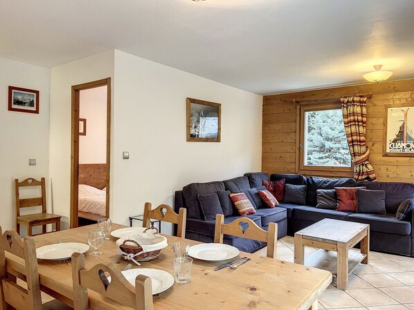 A cozy room with a wooden dining table set for two, a dark blue sofa, and a window with a view of trees.